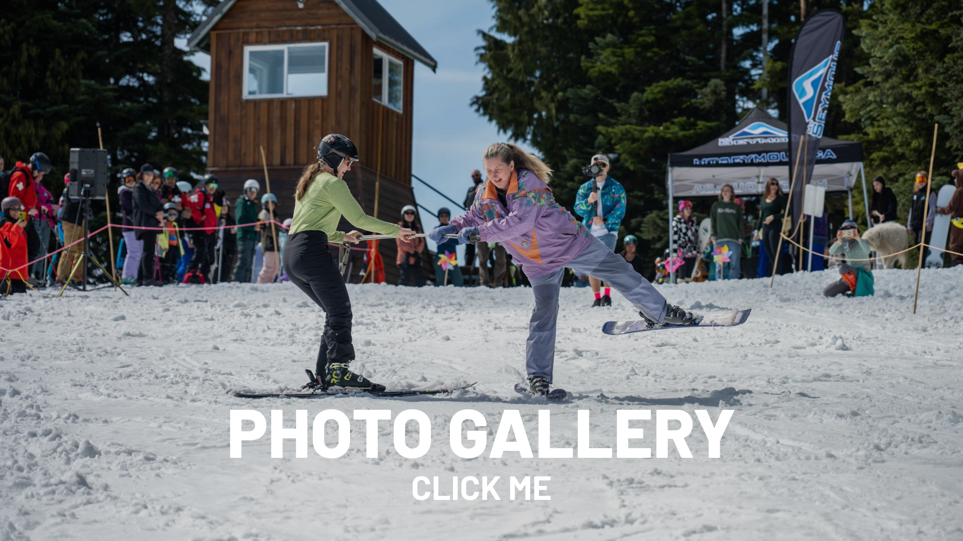 Two people skiing on snow with a crowd and trees in the background.