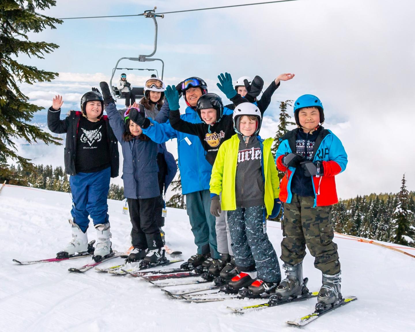 Group of kids in winter gear standing on snowy ski slope, smiling and waving.