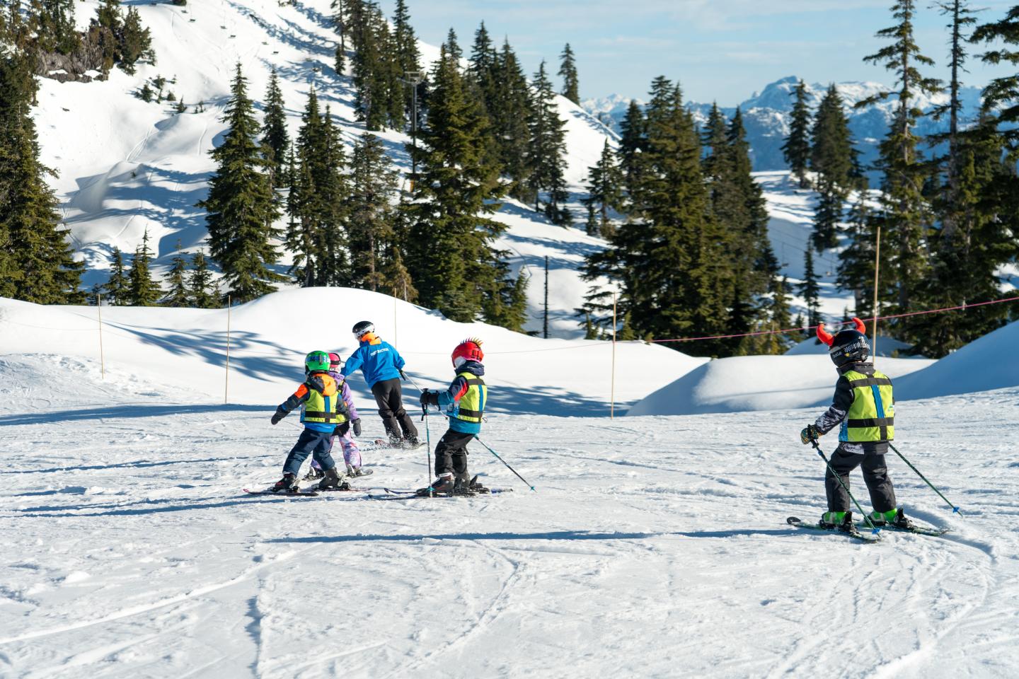 Children skiing on a snowy mountain with instrcutor & evergreen trees.