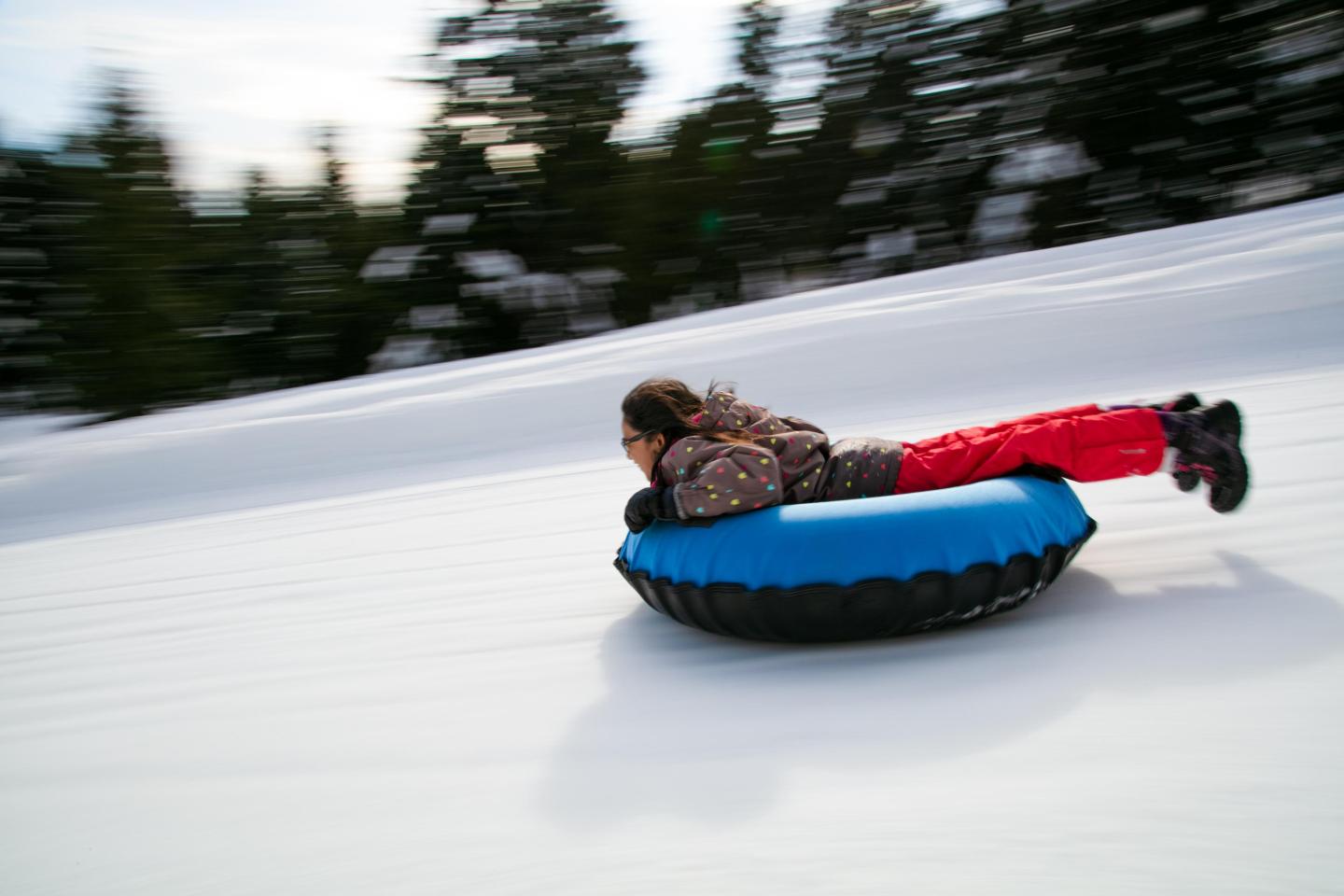 Child sliding down snowy hill on a blue inner tube, wearing red pants.