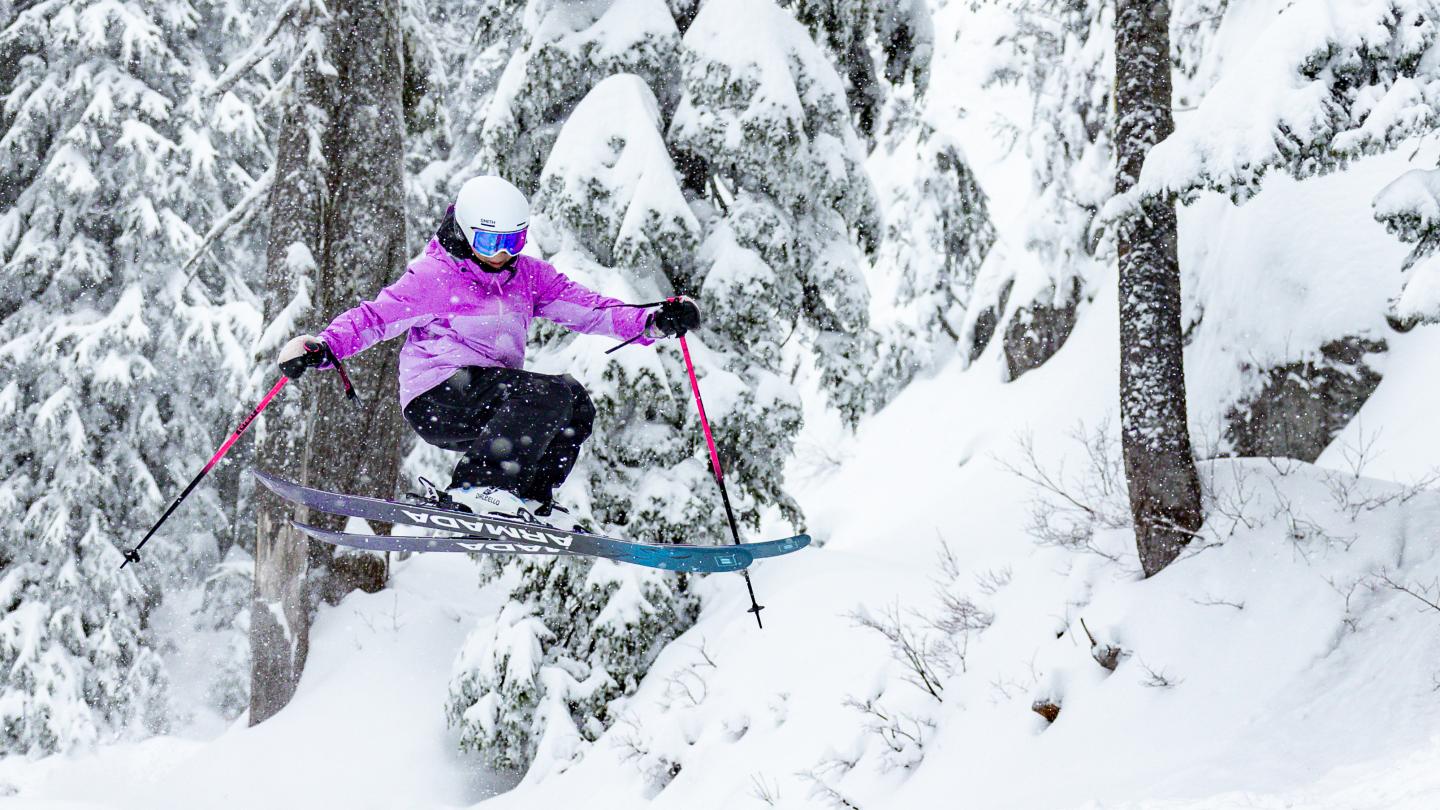Skier in a purple jacket jumps through snowy forest.