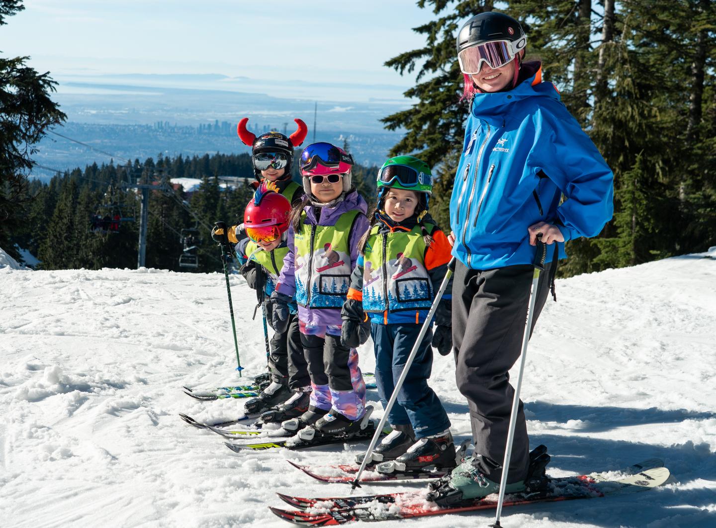 Ski instructor and kids in colorful outfits on snowy slope, trees in background.