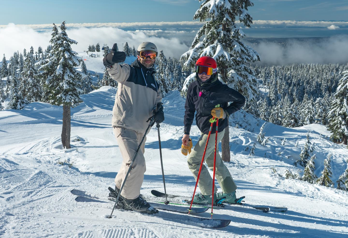 Two people skiing on a snowy mountain, trees and clouds in the background.
