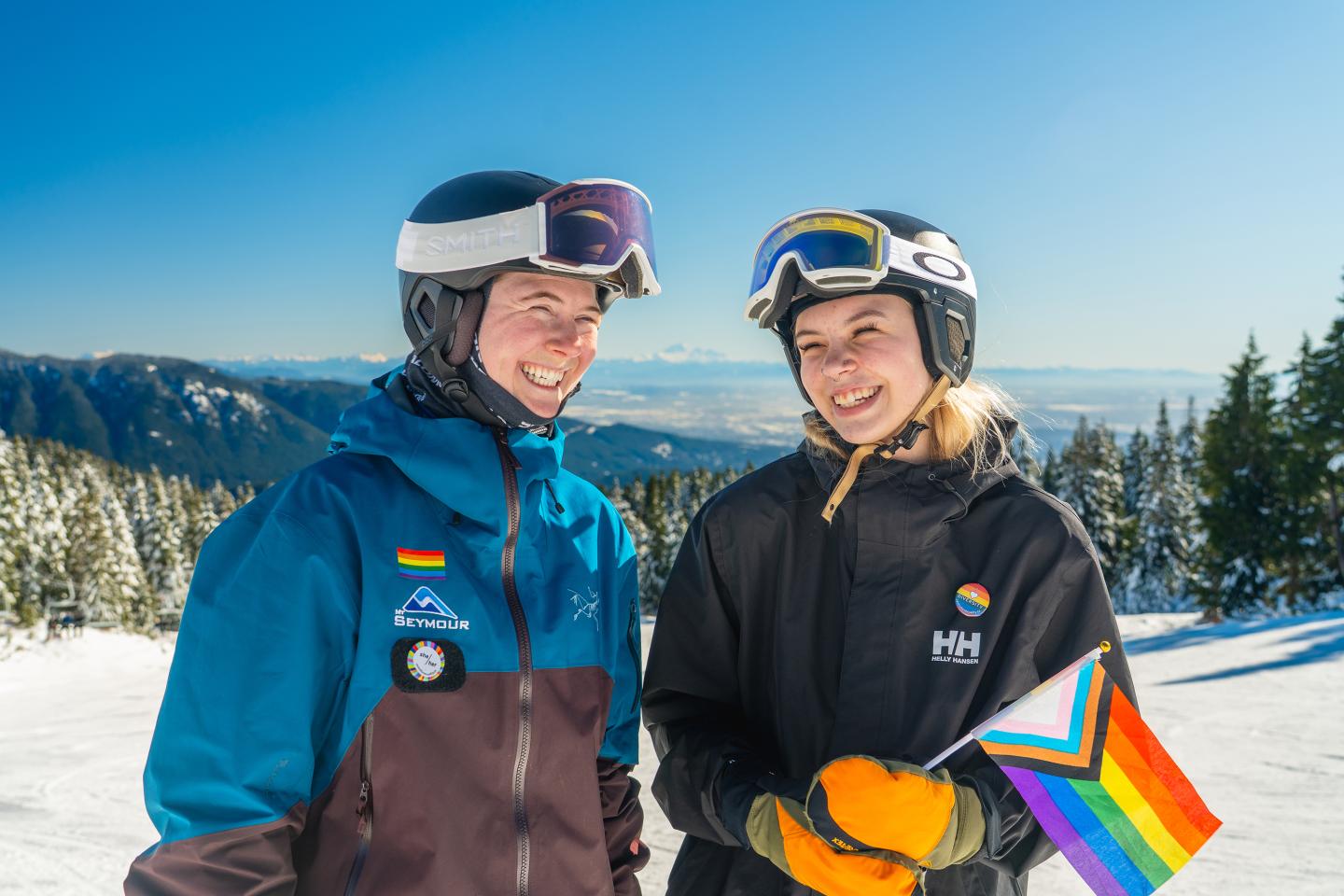 Two skiers in helmets and goggles smiling, holding a rainbow flag on a snowy mountain.