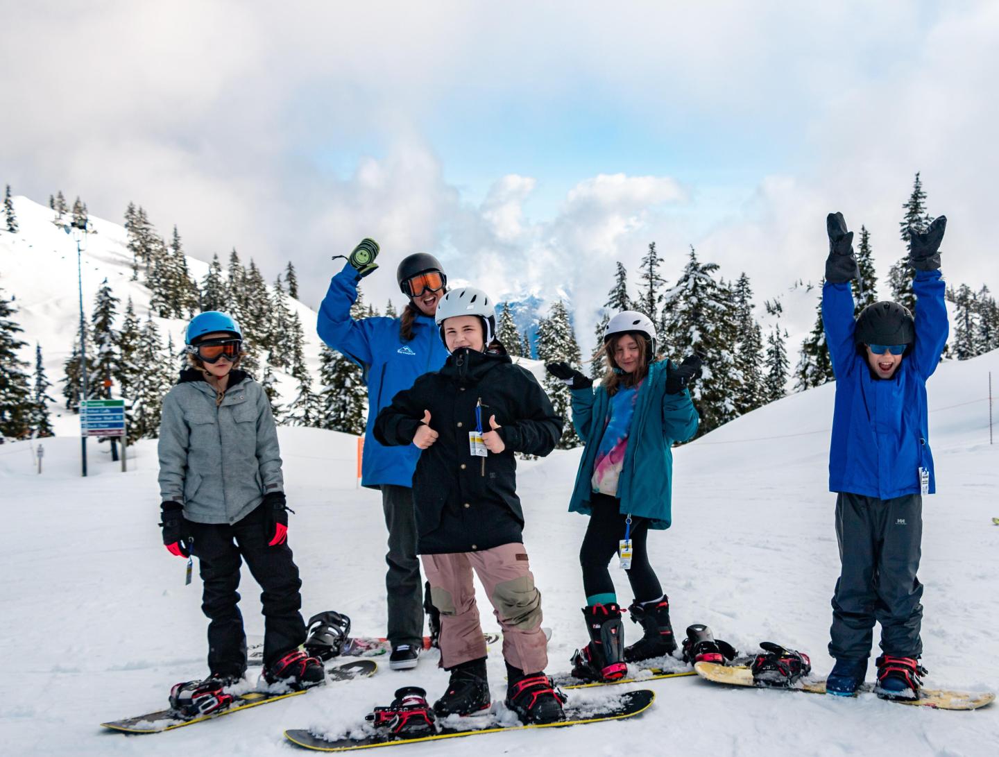 Group of people with snowboards posing on a snowy mountain, trees in the background.