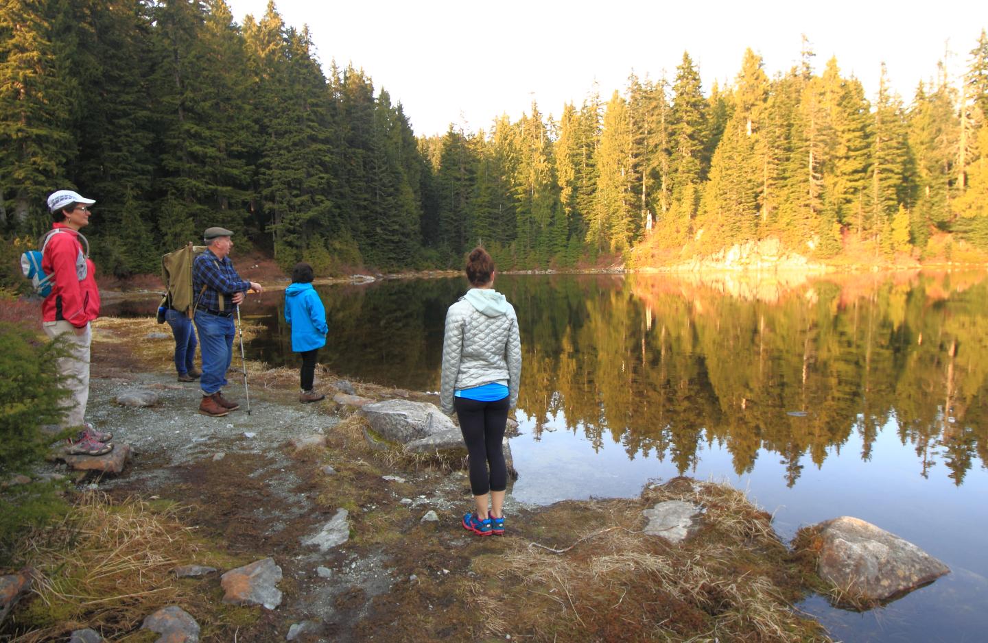 A group enjoys a serene lake surrounded by evergreen trees under golden light.