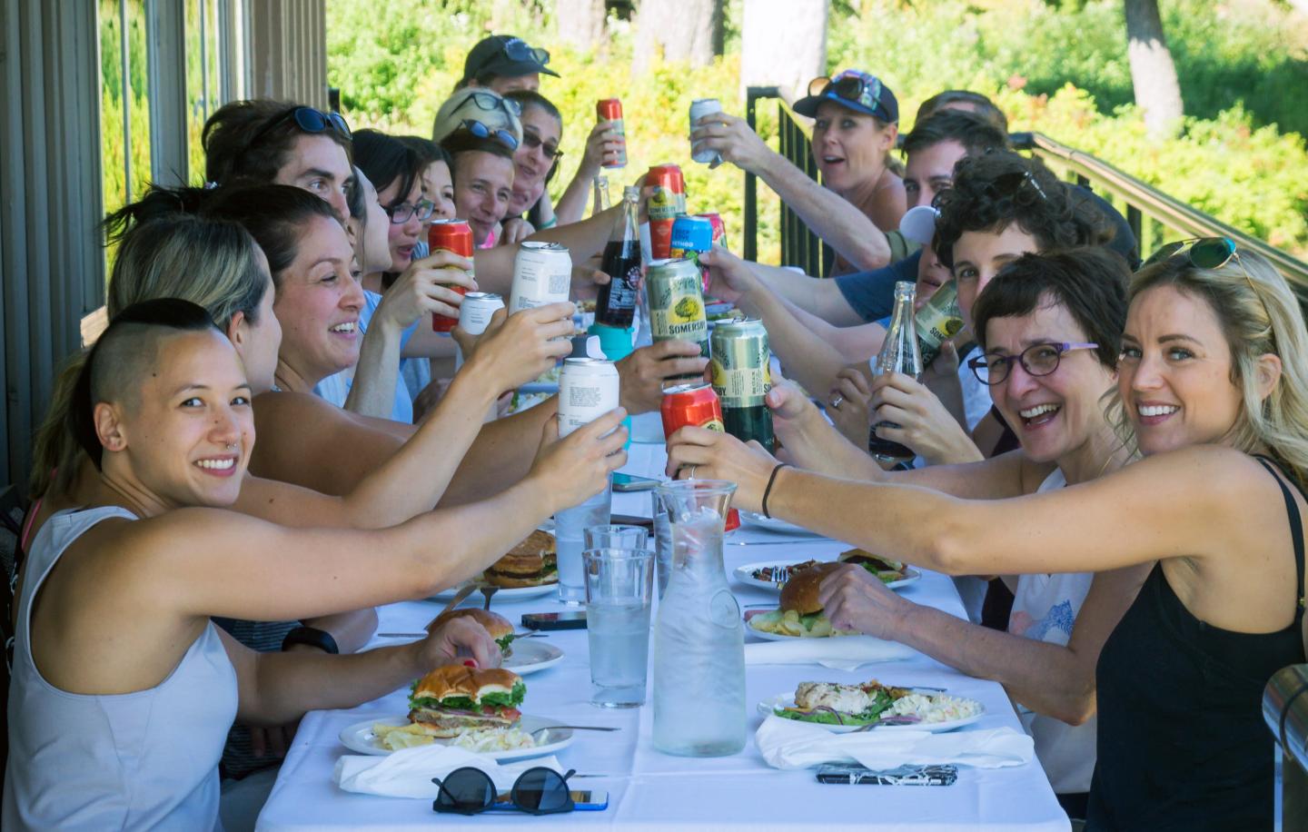 People sitting at a long table, toasting with drinks, smiling outdoors.