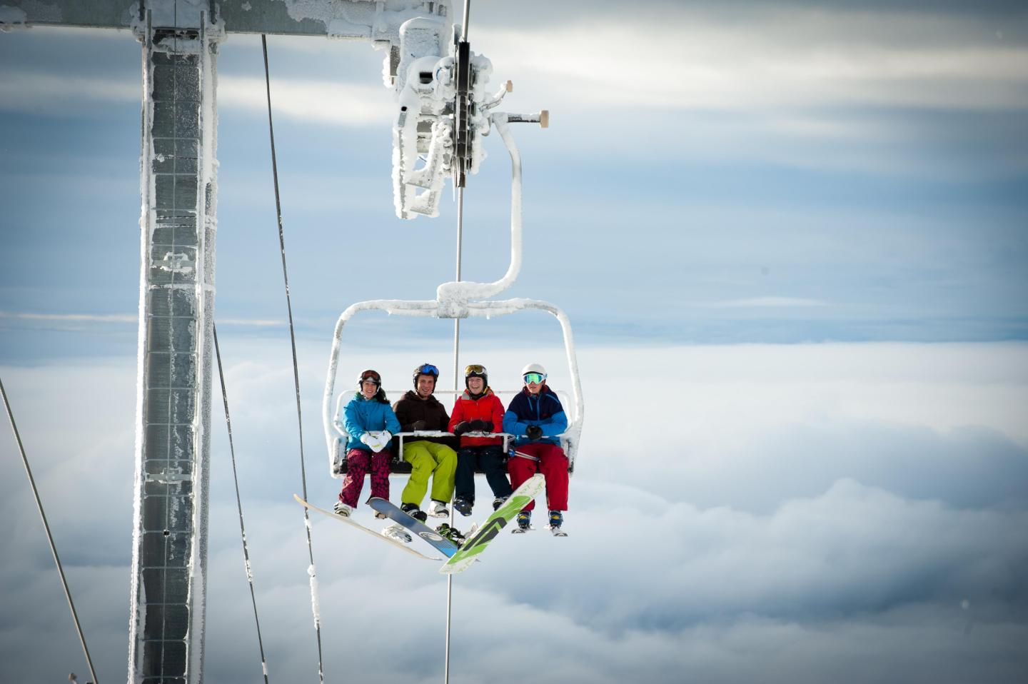 Four people on a ski lift above clouds, wearing colorful ski gear.