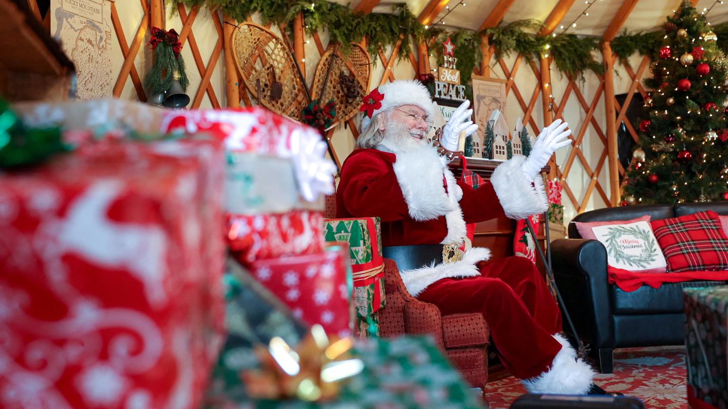 Santa reading a letter beside wrapped gifts and a decorated Christmas tree.