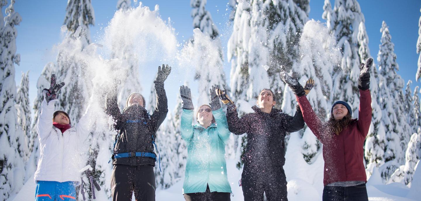 Five people in winter clothing joyfully throwing snow, snowy trees in background.