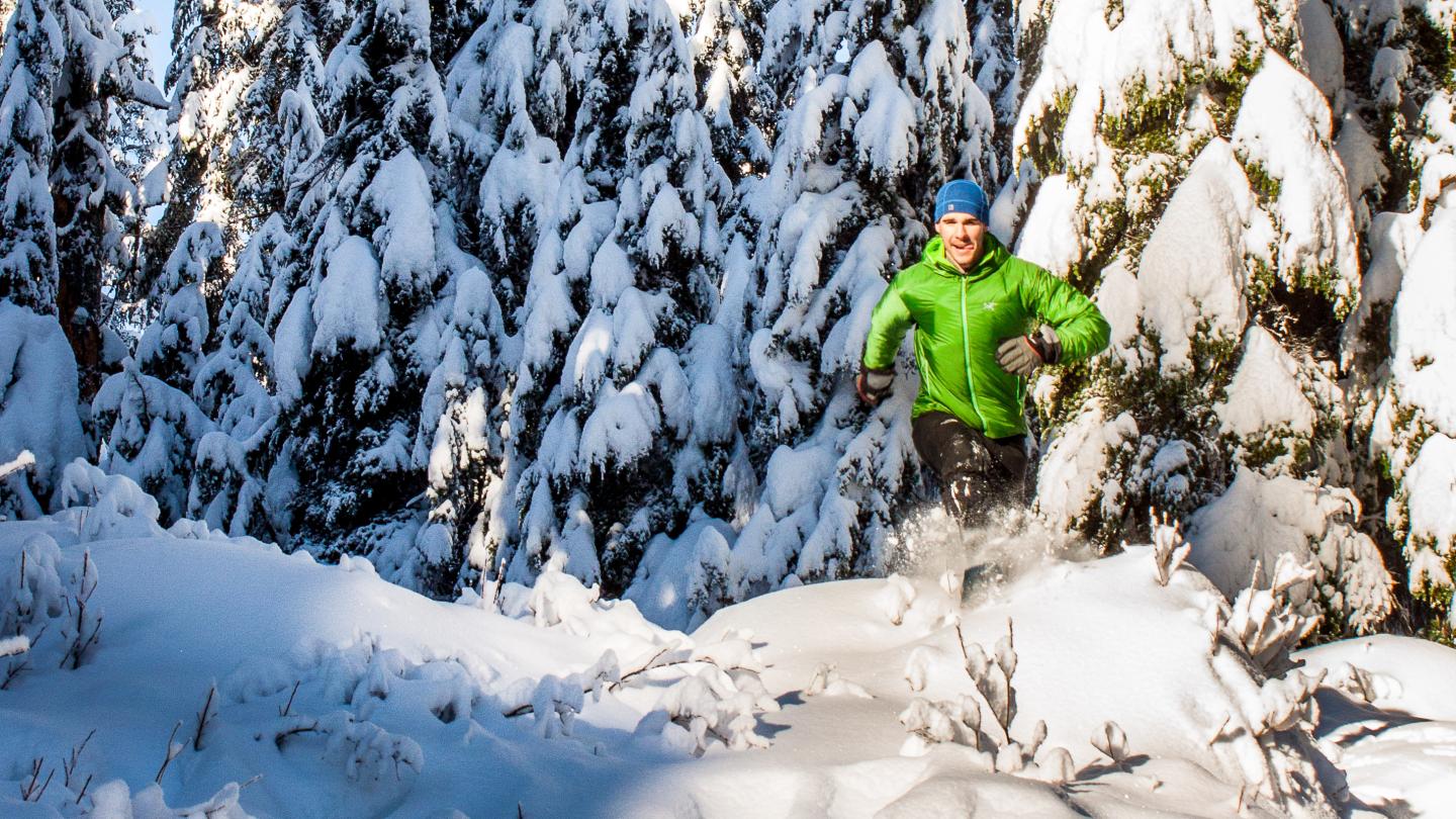 Man jogging in snowy forest, wearing green jacket and blue hat.