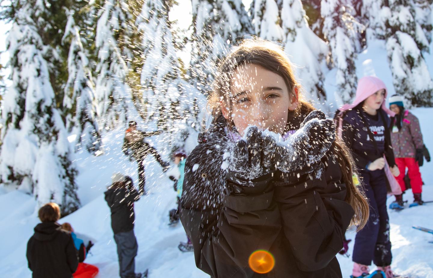 Girl blowing snow in a snowy forest, with kids playing in the background.