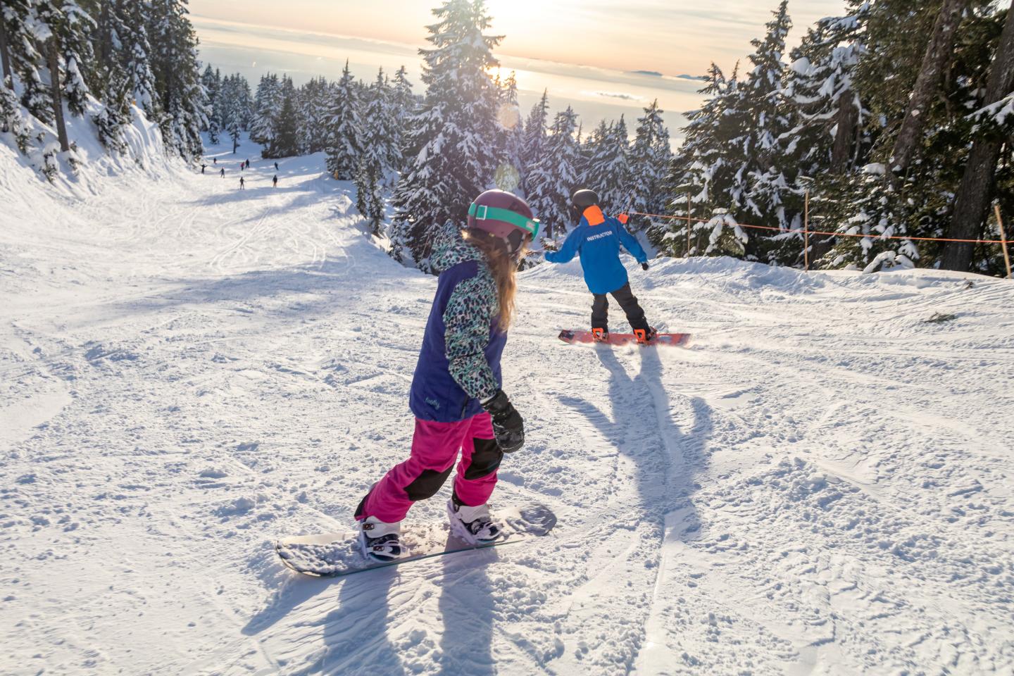 Two children snowboarding down a sunny, tree-lined slope.